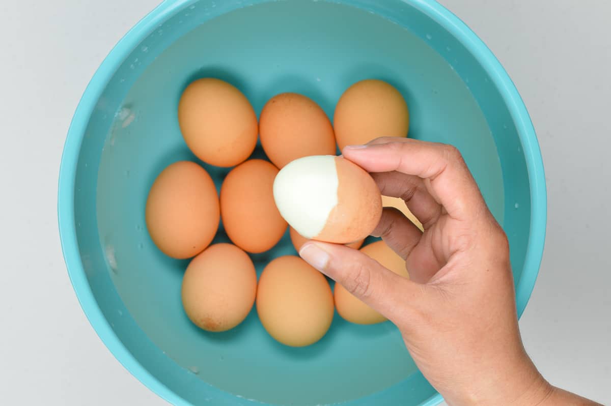 a hand holding a half peeled instant pot hard boiled egg over a bowl of unpeeled boiled eggs in the ice bath