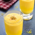 a glass full of mango lassi, garnished with saffron strands and crushed pistachios, with another such glass in the background