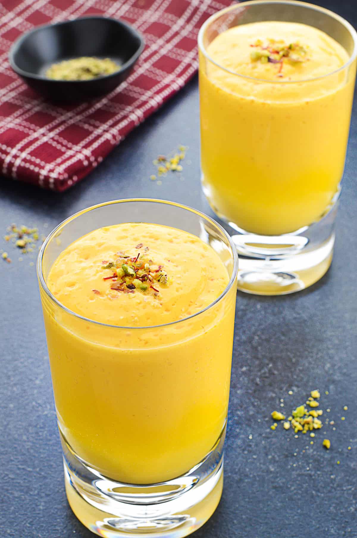 a glass full of mango lassi, garnished with saffron strands and crushed pistachios, with another such glass in the background