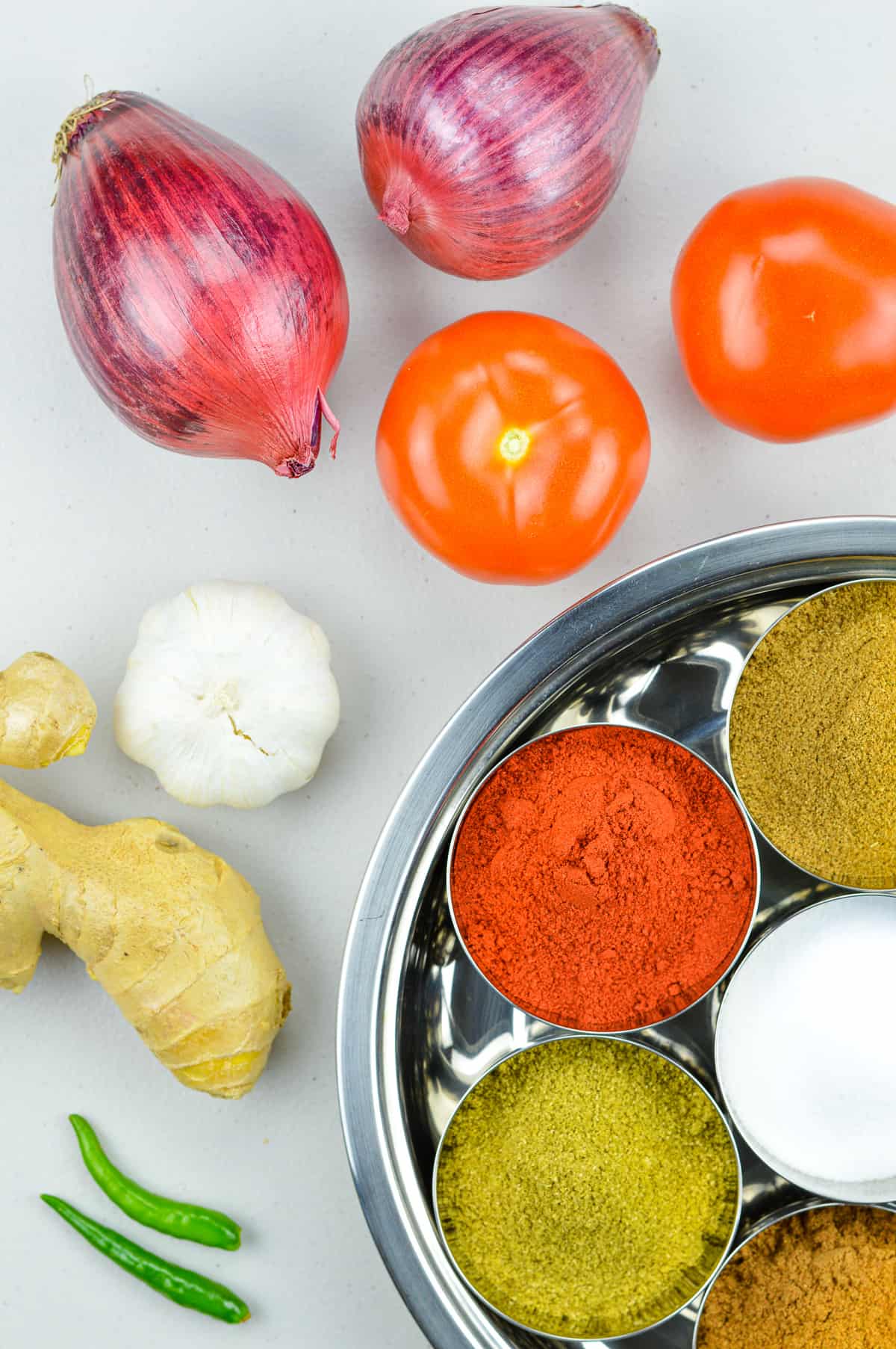 raw ingredients of bhuna masala placed on a white table - including onions, tomatoes, ginger, garlic, chillies, and indian spice powders