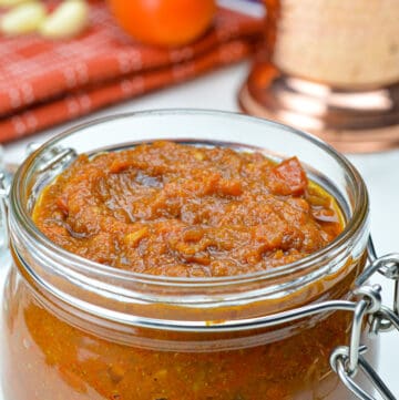 a glass jar filled with bhuna masala curry sauce base, with the raw ingredients (onion, tomato, ginger & garlic) in the background