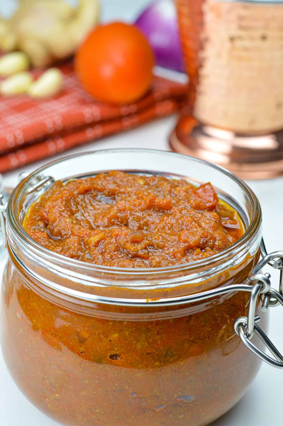 a glass jar filled with bhuna masala curry sauce base, with the raw ingredients (onion, tomato, ginger & garlic) in the background