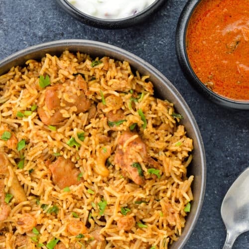 boneless chicken biryani served in a bowl, with yogurt and spicy curry on the side