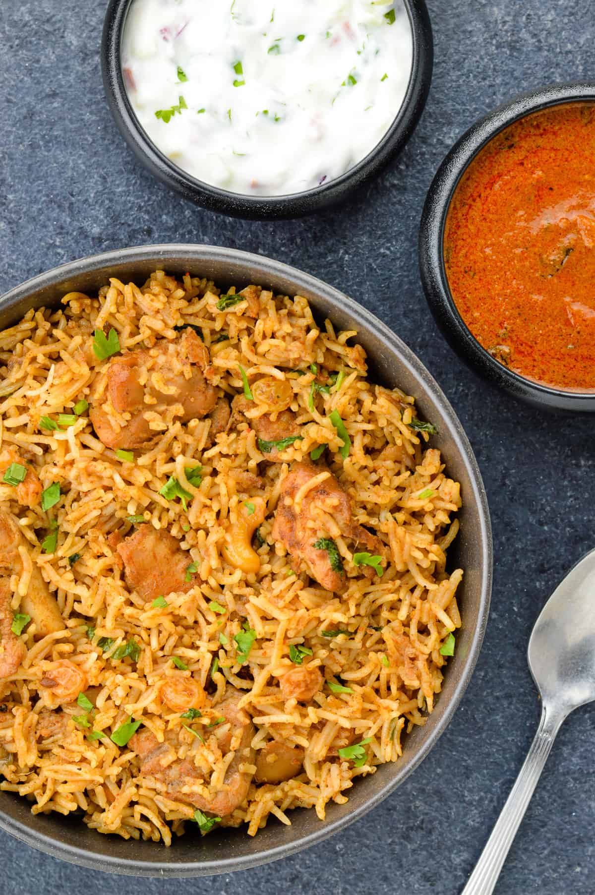 boneless chicken biryani served in a bowl, with yogurt and spicy curry on the side