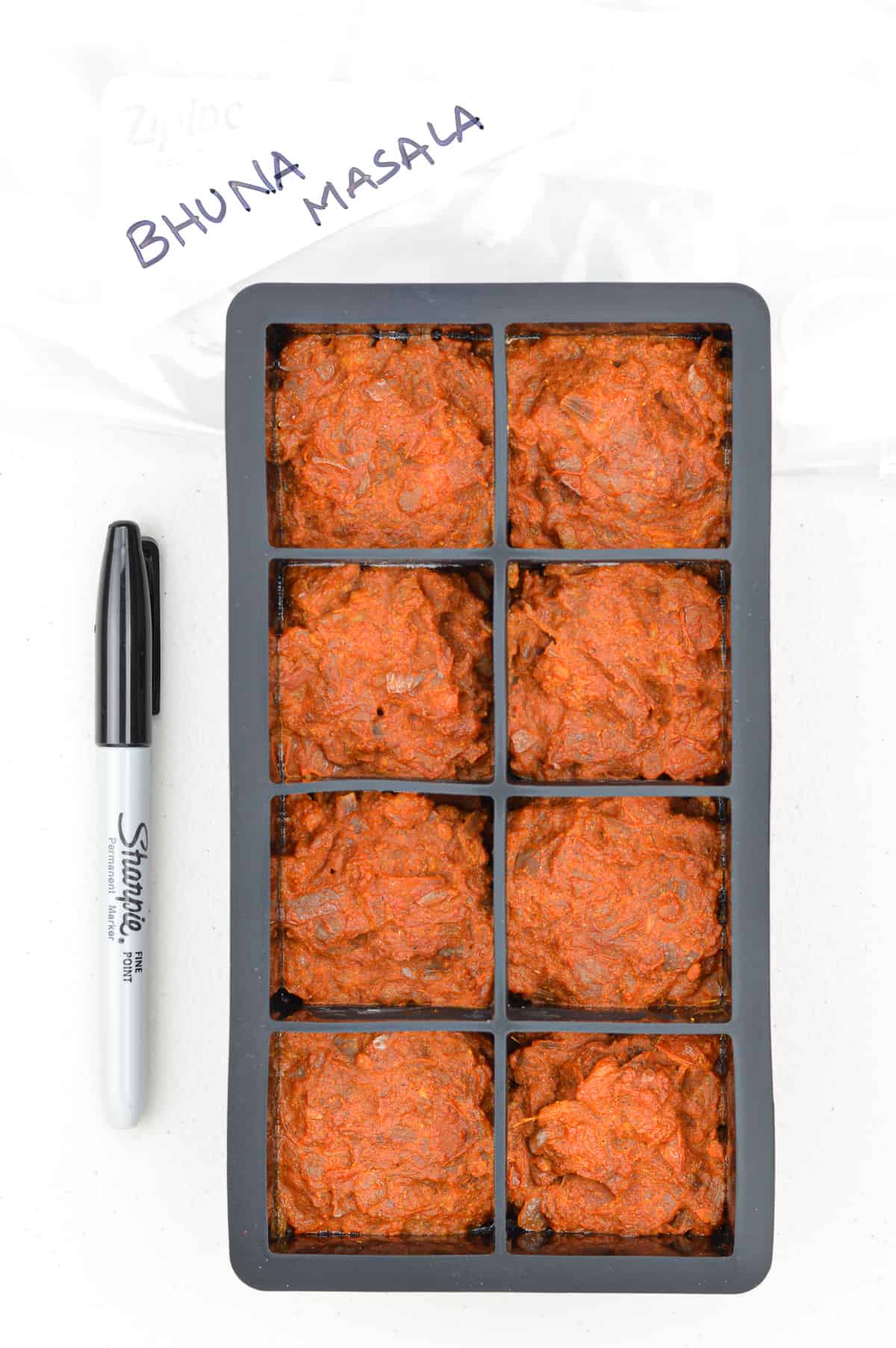 frozen cubes of bhuna masala curry sauce base in a black ice cube tray, with a marker pen & ziplock bag on the side