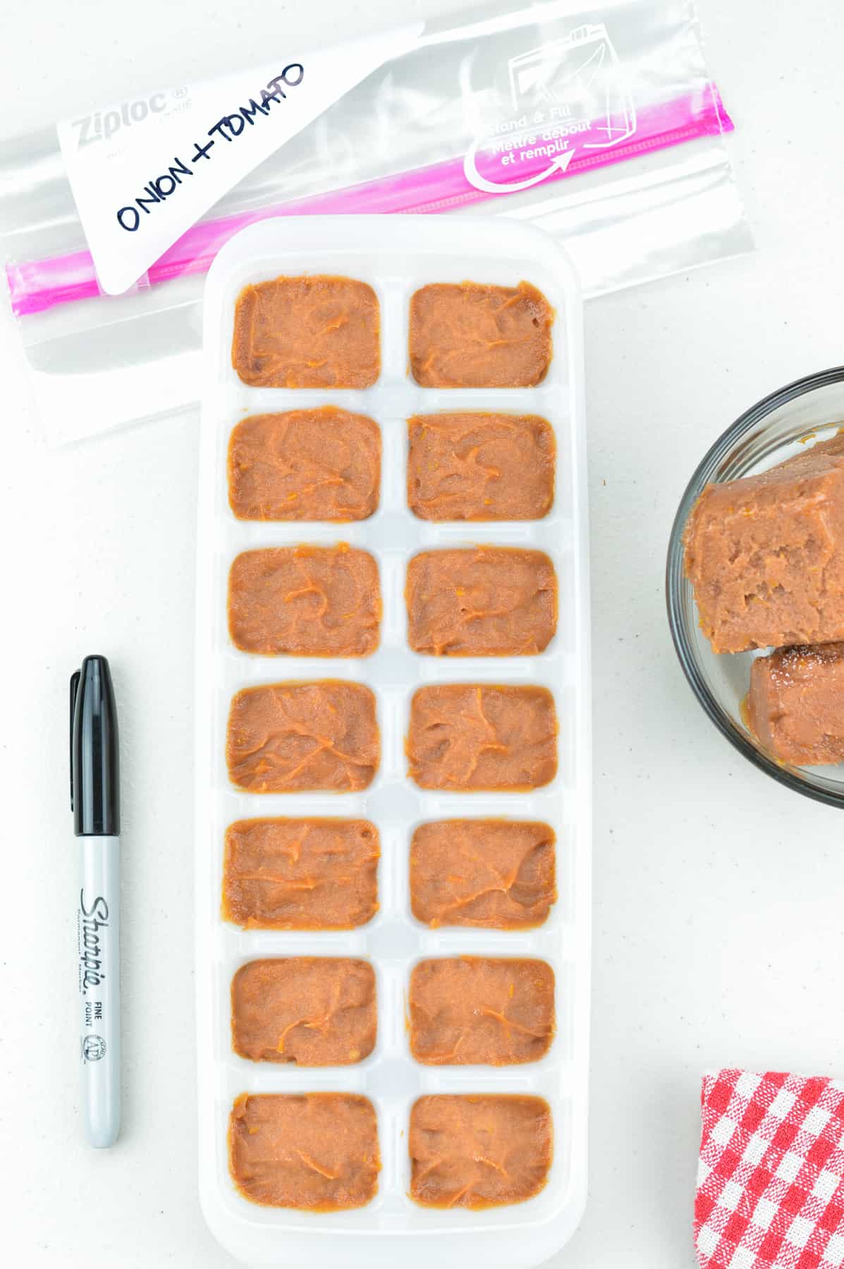 frozen onion tomato masala paste in an ice cube tray, with some frozen masala paste cubes in a bowl on one side, and a marker pen & ziplock bag on the other side