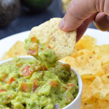 a hand dipping into a bowl full of guacamole with a chip, with more chips on the side, in the background, along with 2 avocados