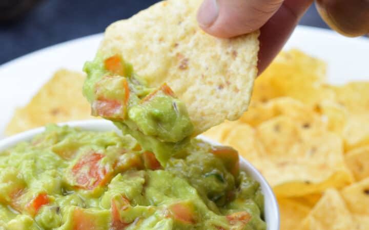 a hand dipping into a bowl full of guacamole with a chip, with more chips on the side, in the background, along with 2 avocados