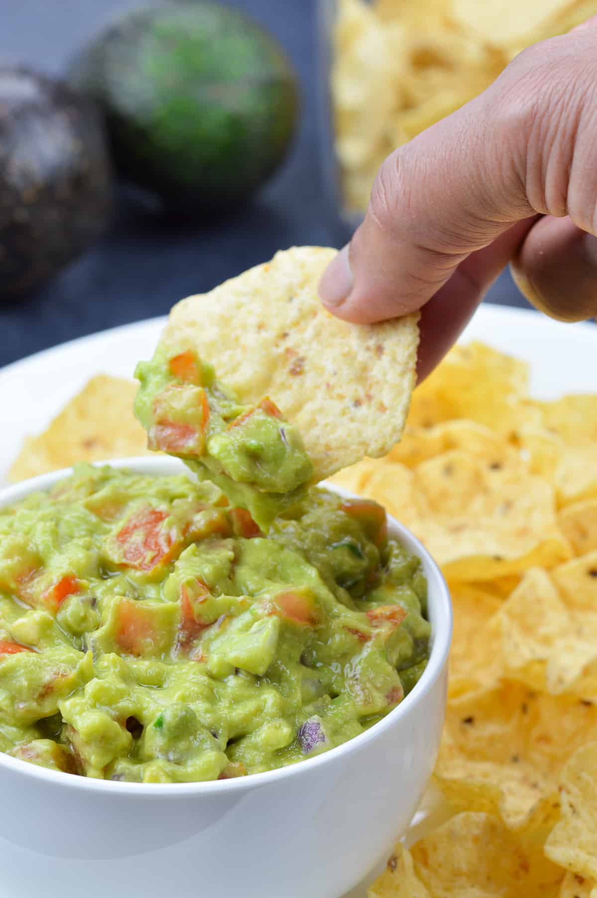 a hand dipping into a bowl full of guacamole with a chip, with more chips on the side, in the background, along with 2 avocados