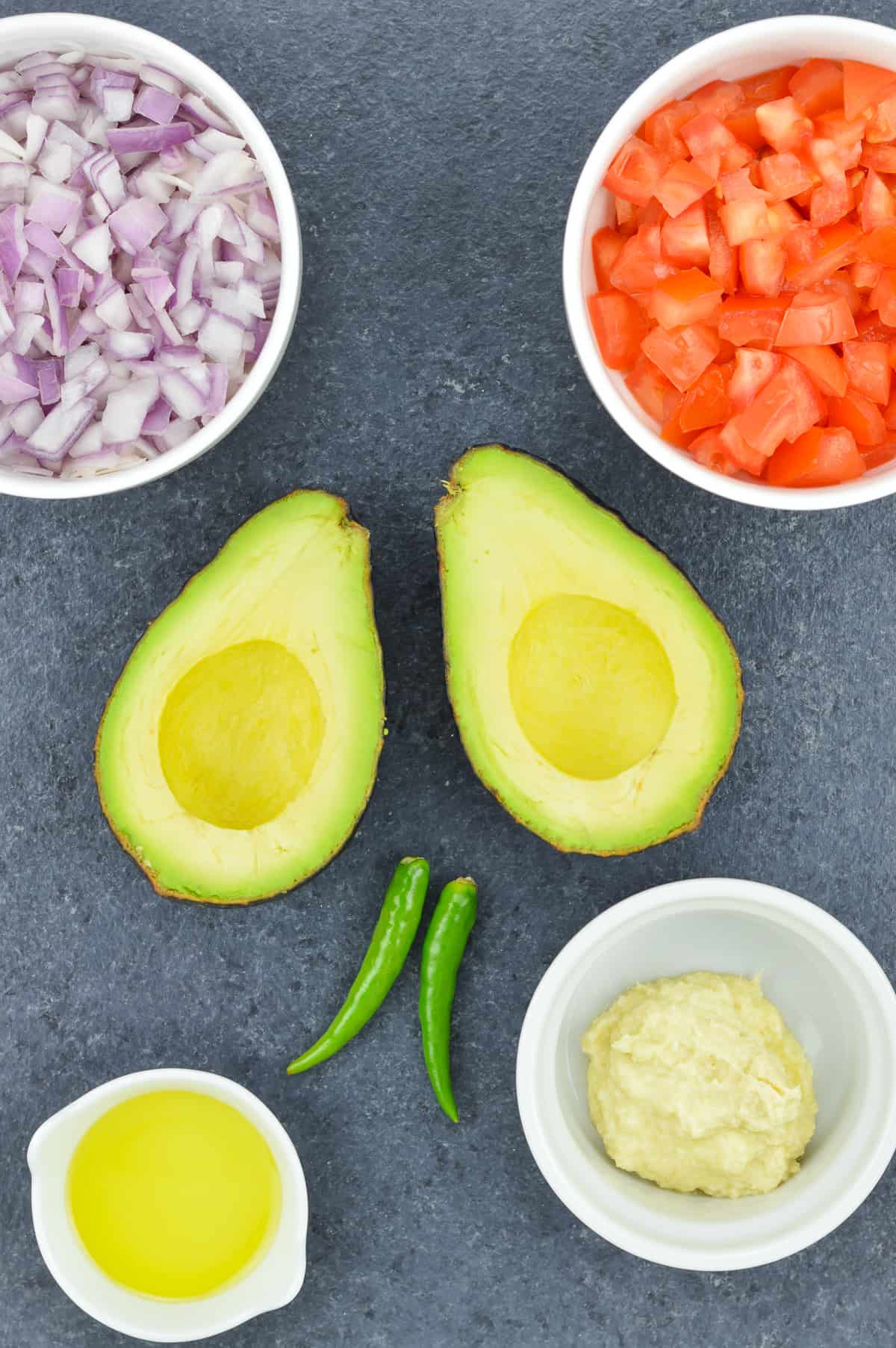 ingredients to make indian style guacamole placed on a dark surface - one avocado cut vertically into two pieces, and placed face up, along with bowl of chopped onions, chopped tomatoes, ginger garlic paste, extra virgin olive oil, and 2 green chillies