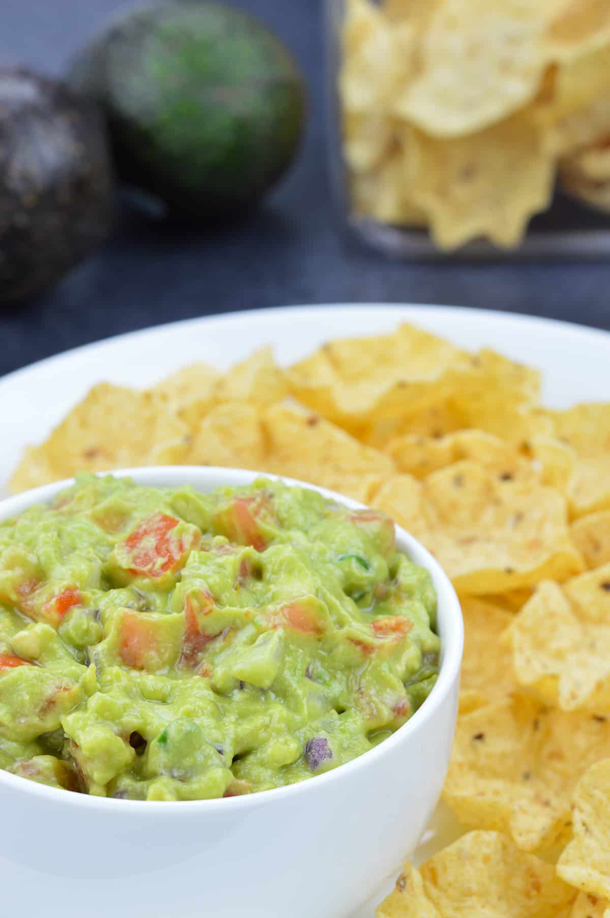 a bowl full of guacamole, with tostitos chips on the side, and in the background, along with 2 avocados