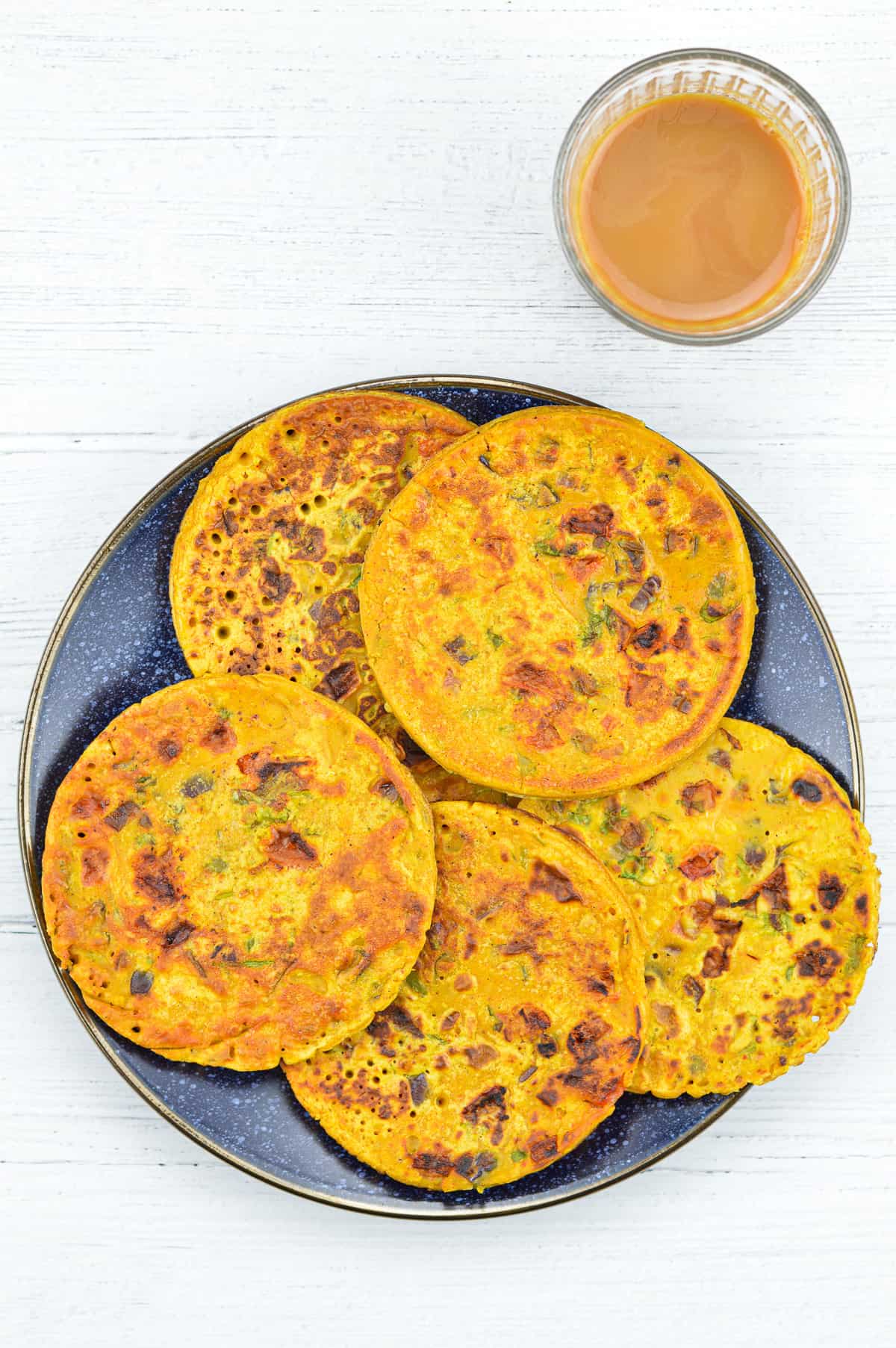 5 small besan chillas on a plate, with a glass of tea on the side