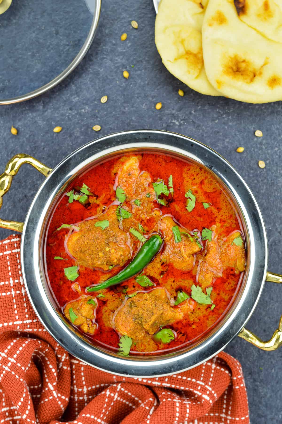 a serving kadai full of kolhapuri chicken curry, with a plate of naan and kitchen towel on either side