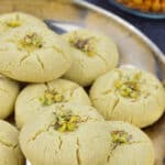 a plate of nankhatai biscuits garnished with pistachios and saffron strands, with indian snacks in the background