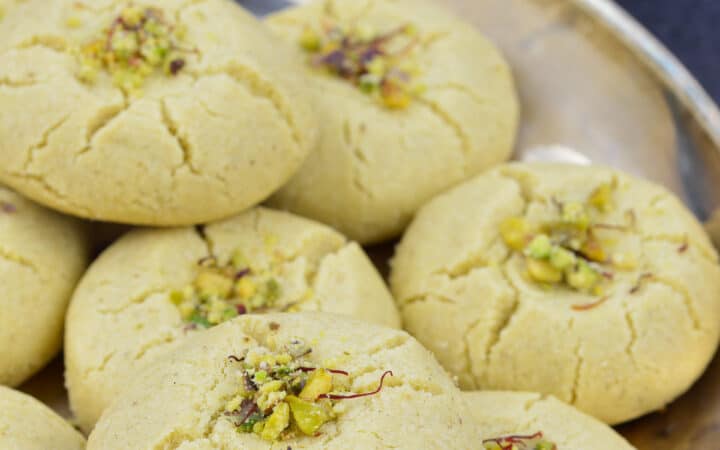 a plate of nankhatai biscuits garnished with pistachios and saffron strands, with indian snacks in the background