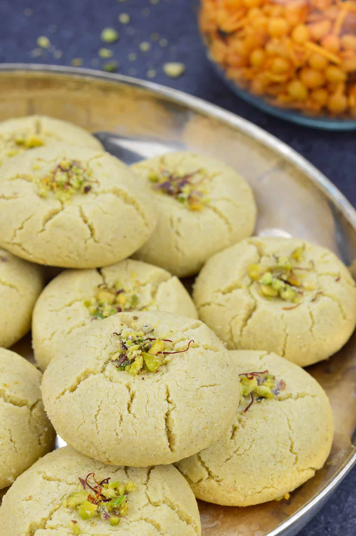 a plate of nankhatai biscuits garnished with pistachios and saffron strands, with indian snacks in the background.