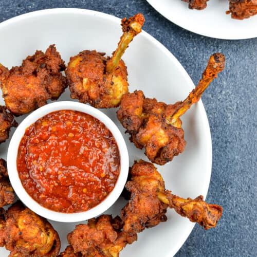 chicken lollipops placed around a small bowl of schezwan chutney in a white plate, with a few more lollipops visible in the background