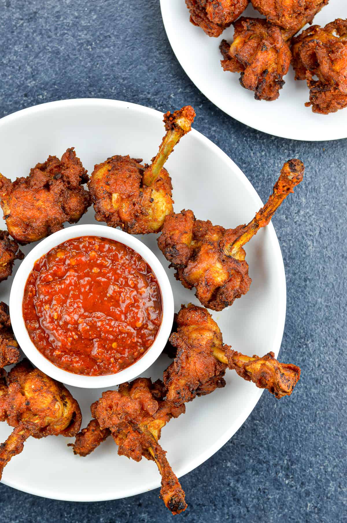 chicken lollipops placed around a small bowl of schezwan chutney in a white plate, with a few more lollipops visible in the background