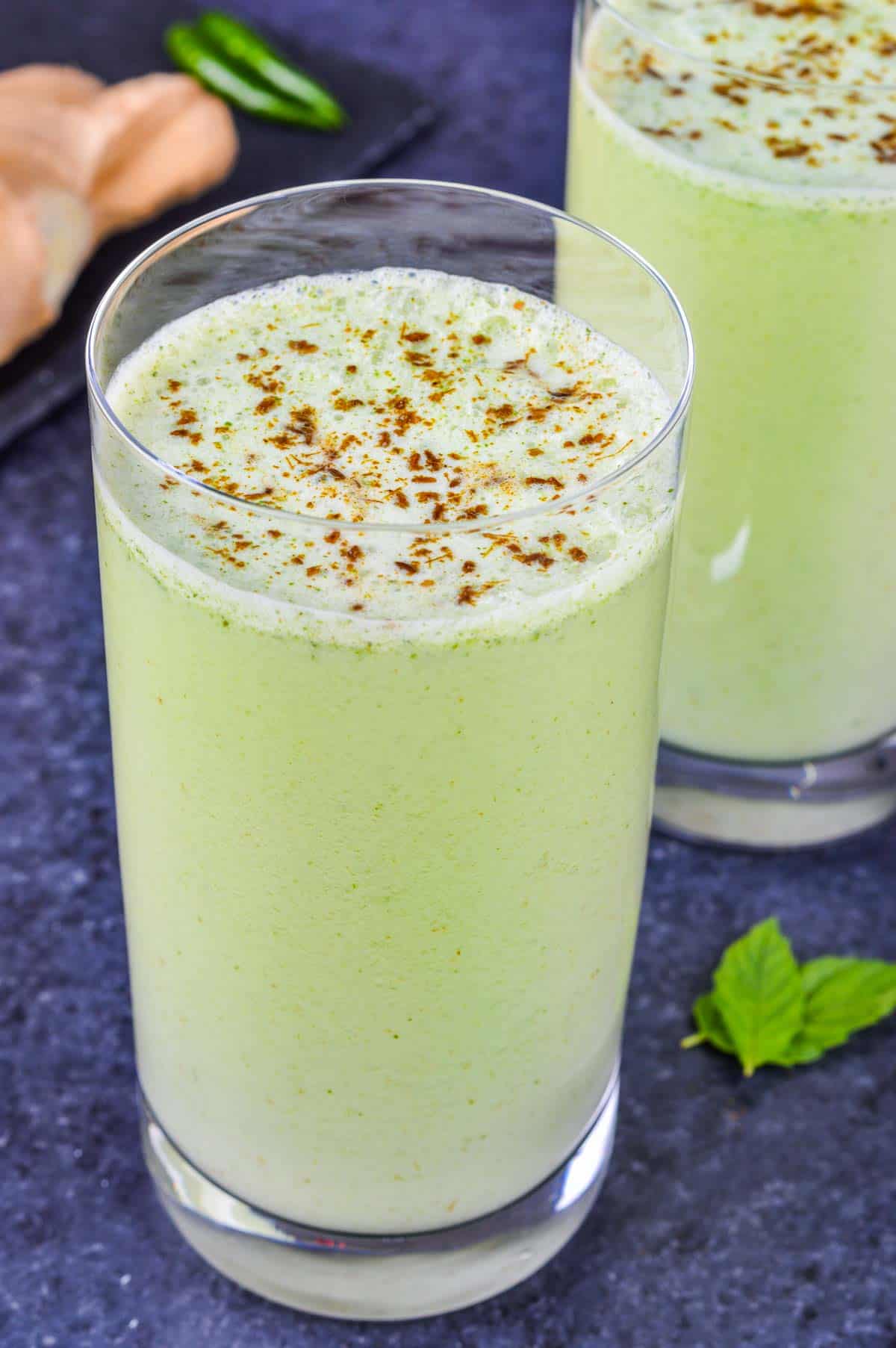 close up shot of a glass full of masala chaas, with another glass of chaas in the background, along with some whole ginger, chillies, and mint leaves on the side.