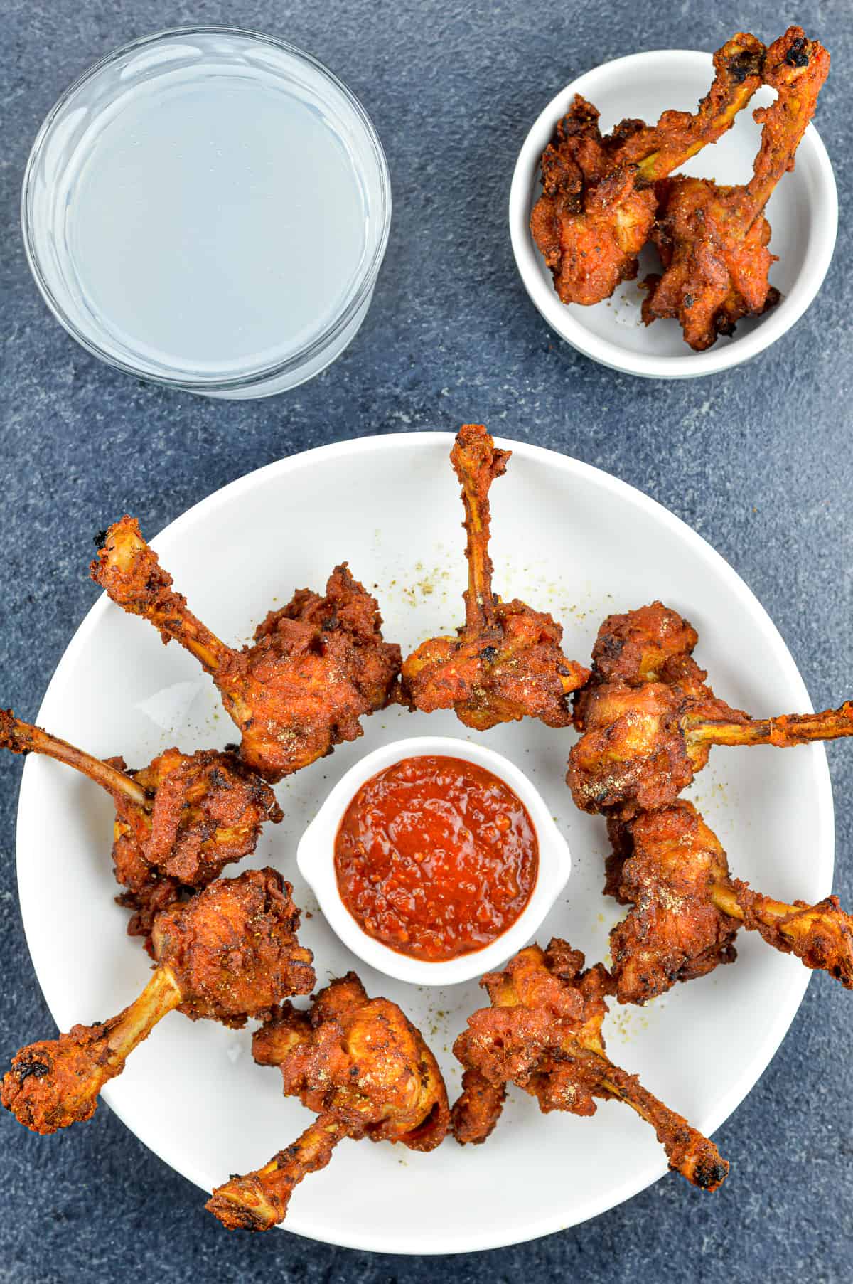 chicken lollipops placed around a small bowl of schezwan chutney in a white plate, served along with a drink, and a few extra lollipops on the side