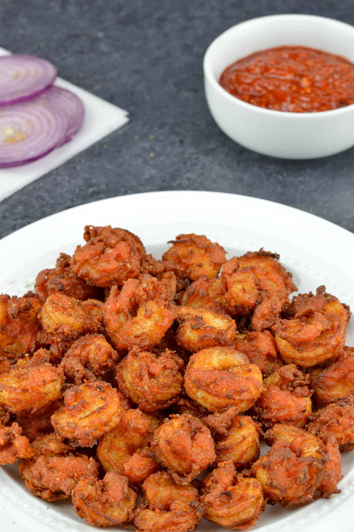 crispy fried prawns koliwada sprinkled with chaat masala, served in a white plate, along with schezwan chutney, and sliced onions in the background