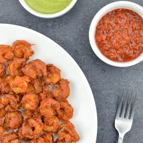 mumbai style prawns koliwada sprinkled with chaat masala, served in a white plate, along with a fork, green chutney, and schezwan chutney on the side