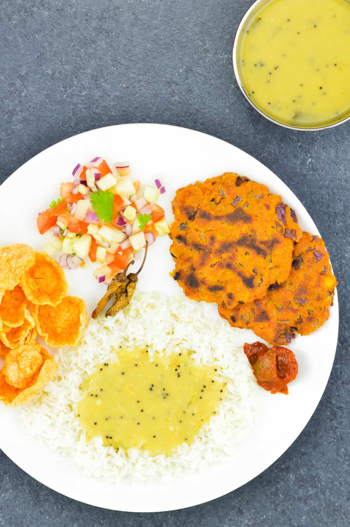  un shot de riz et de dal en haut servi dans une assiette blanche, accompagné d'un chana dal chilla, de quelques croquants, de légumes hachés et de cornichons indiens, avec un katori de dal supplémentaire sur le côté