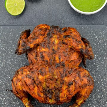 a whole chicken tandoori placed on a black cutting board, with a bowl of green chutney and a couple of lemon wedges on the side