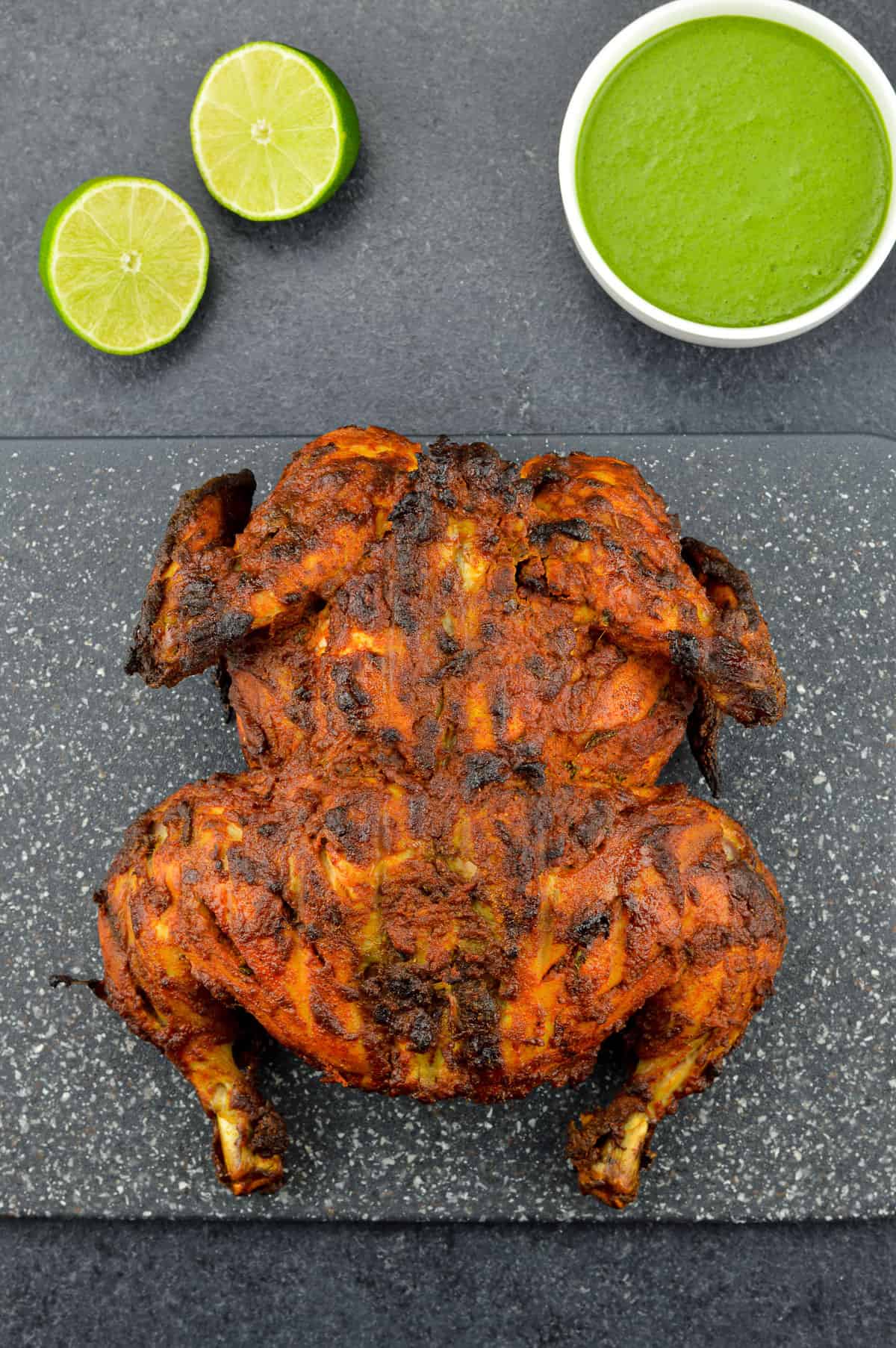 a whole chicken tandoori placed on a black cutting board, with a bowl of green chutney and a couple of lemon wedges on the side