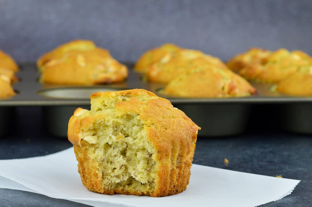 a half eaten white chocolate chip banana muffin placed in front of a tray full of more freshly baked muffins