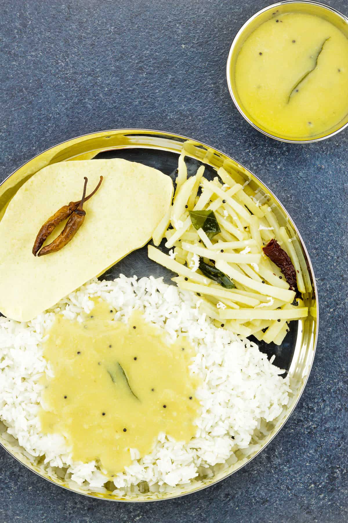 top shot of a konkani thali with dalitoy, rice, batate upkari, and papad, with a small bowl of additional dalitoy on the side