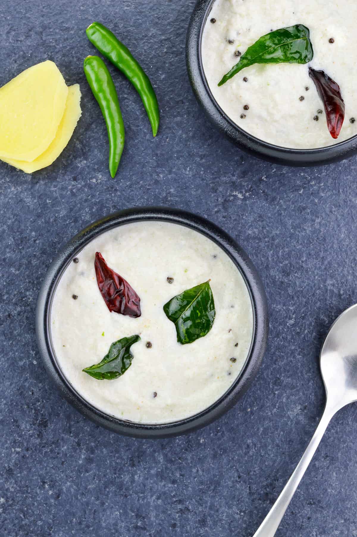 top shot of two bowls of white coconut chutney, with a spoon, few green chillies & some ginger on the side.