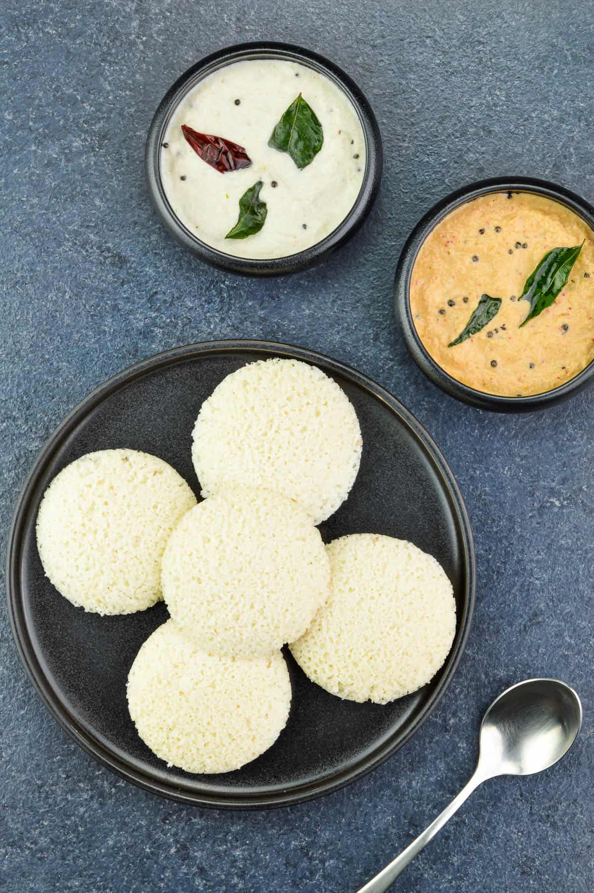 a plate full of steamed idlis, served with 2 small bowls full of 2 varieties of coconut chutney on the side.