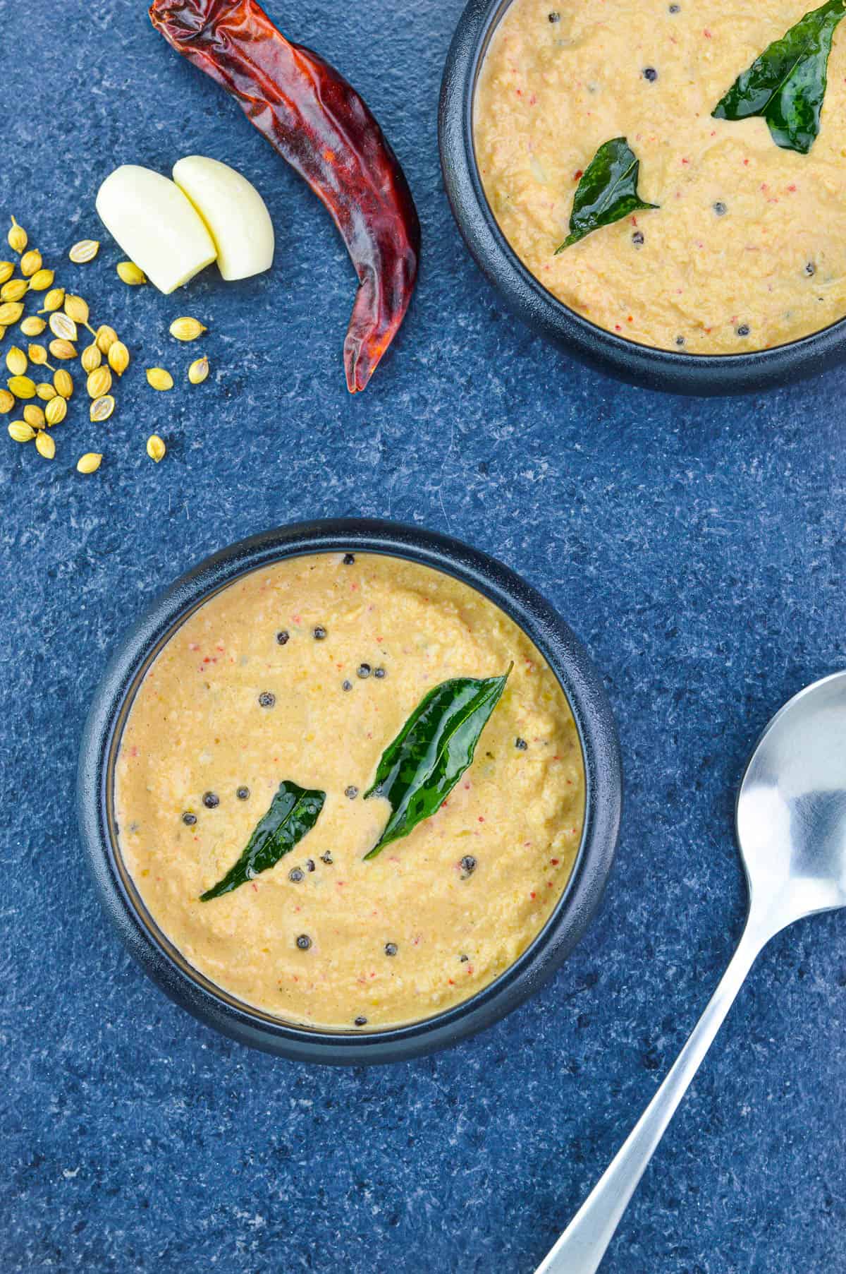 top shot of two bowls of red coconut chutney, along with a spoon, red chilli, few garlic cloves & coriander seeds on the side.