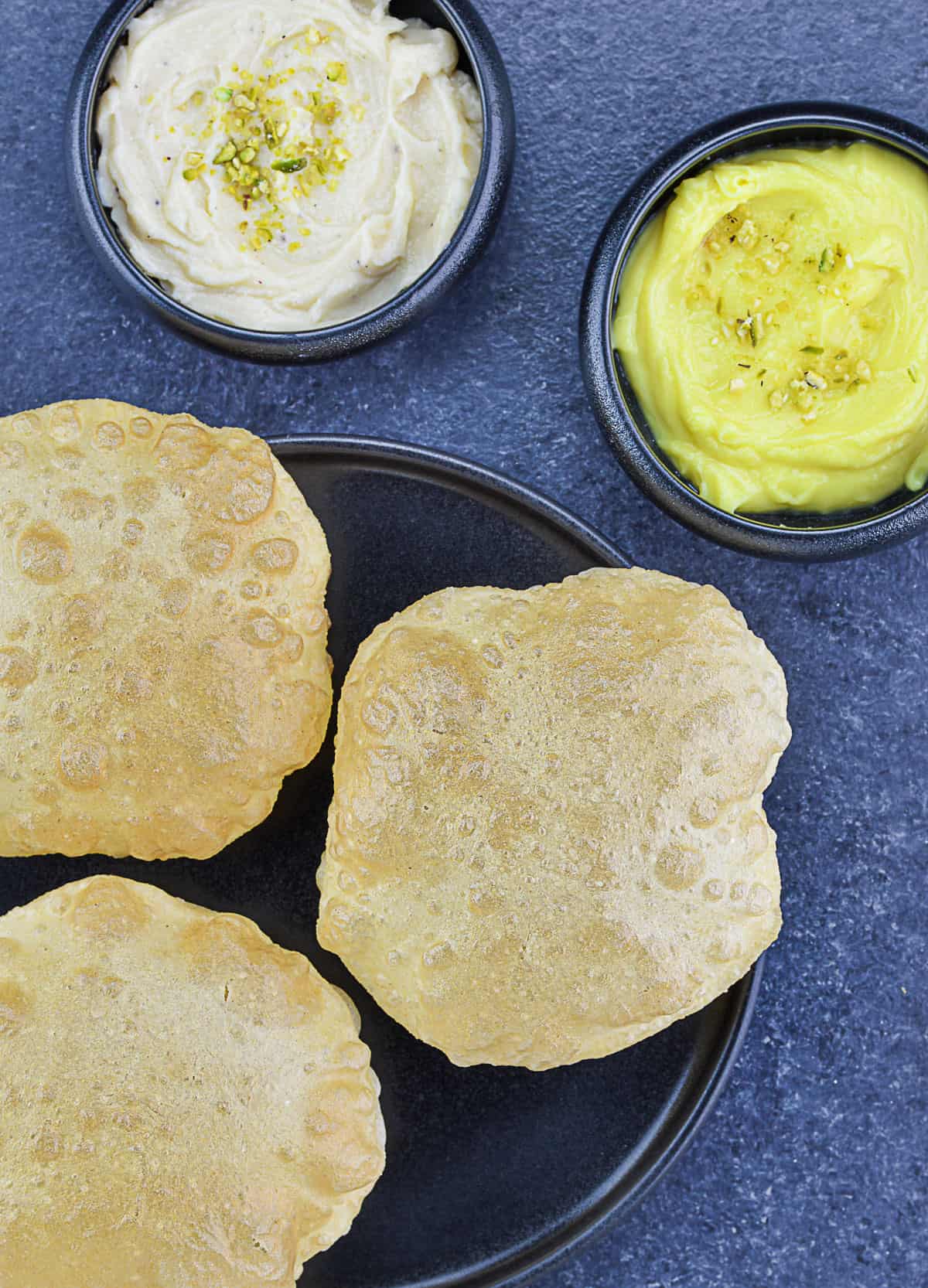 a plate full of puffed puris, served with elaichi shrikhand and pineapple shrikhand on the side