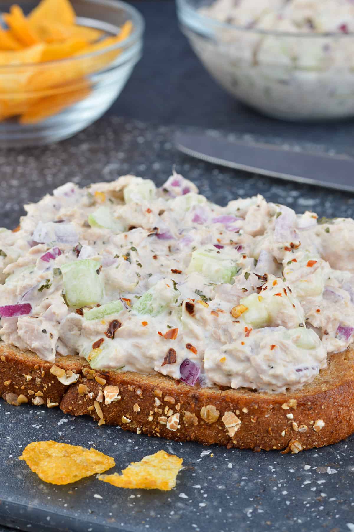 a close up shot of an open faced tuna salad sandwich, with more sandwich filling, and chips in the background