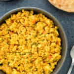 top shot of a bowl full of paneer bhurji, with a roti on the side, along with a spoon & some sliced onions