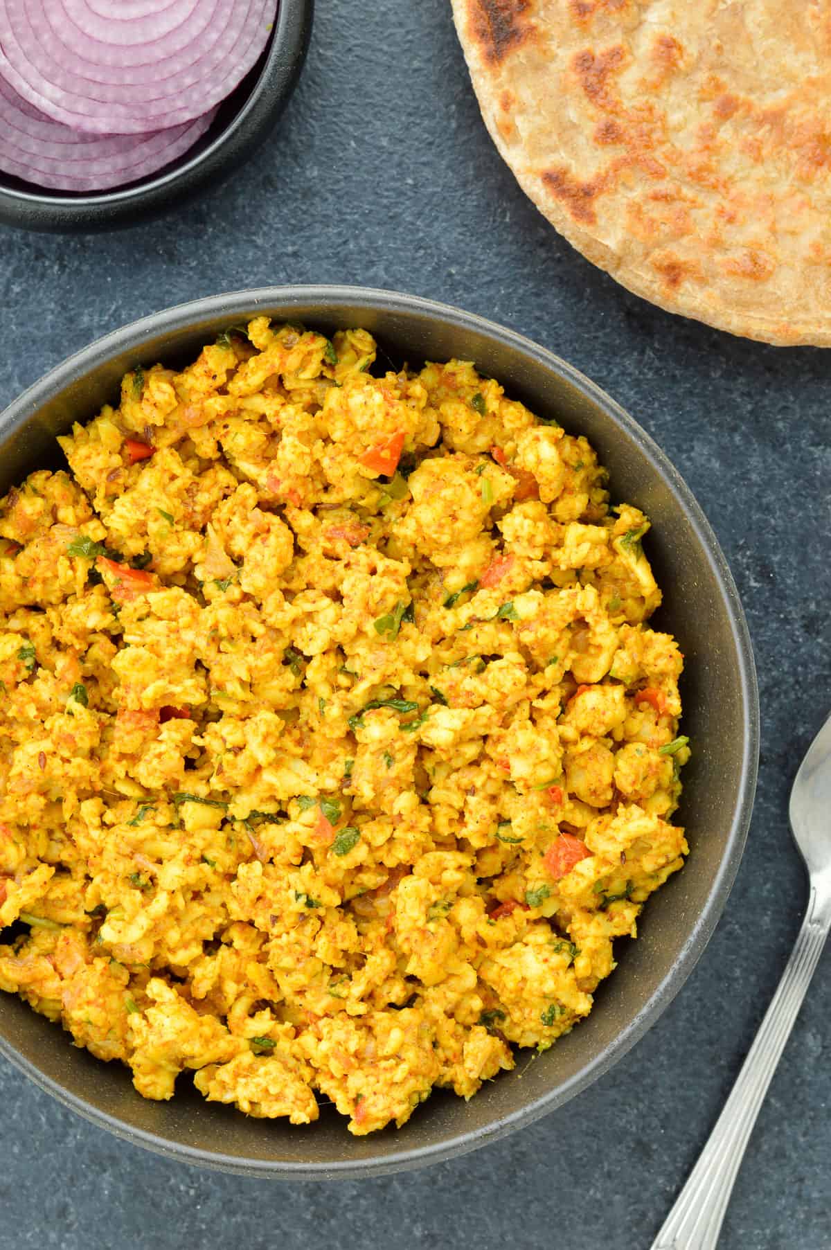 top shot of a bowl full of paneer bhurji, with a roti on the side, along with a spoon & some sliced onions