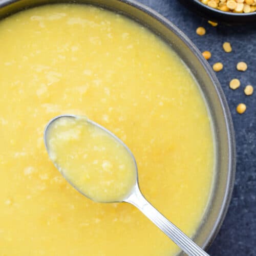 a spoon full of plain boiled yellow dal, held above a bowl full of boiled dal, with a small bowl of raw toor and yellow moong dal in the background