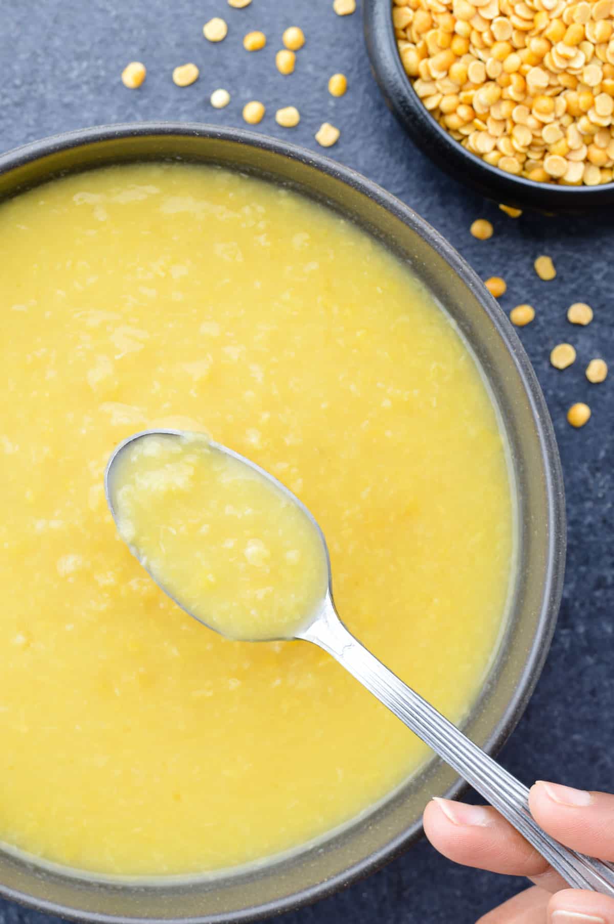 a spoon full of plain boiled yellow dal, held above a bowl full of boiled dal, with a small bowl of raw toor and yellow moong dal in the background