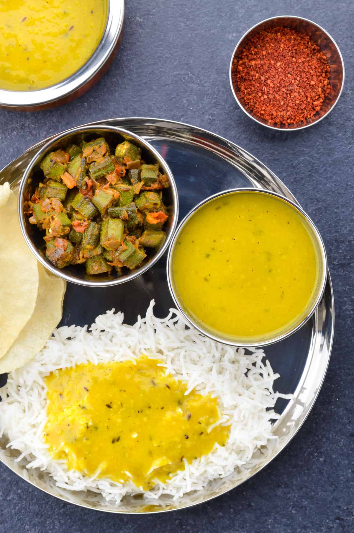 top shot of an indian thali with dal, rice, bhindi sabzi, and papad, along with a small bowl of dry chutney, and a pot of dal on the side.
