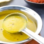 close up of a pot of dal, and a ladle taking some dal from the pot, with a small bowl of chutney and a papad in the background.