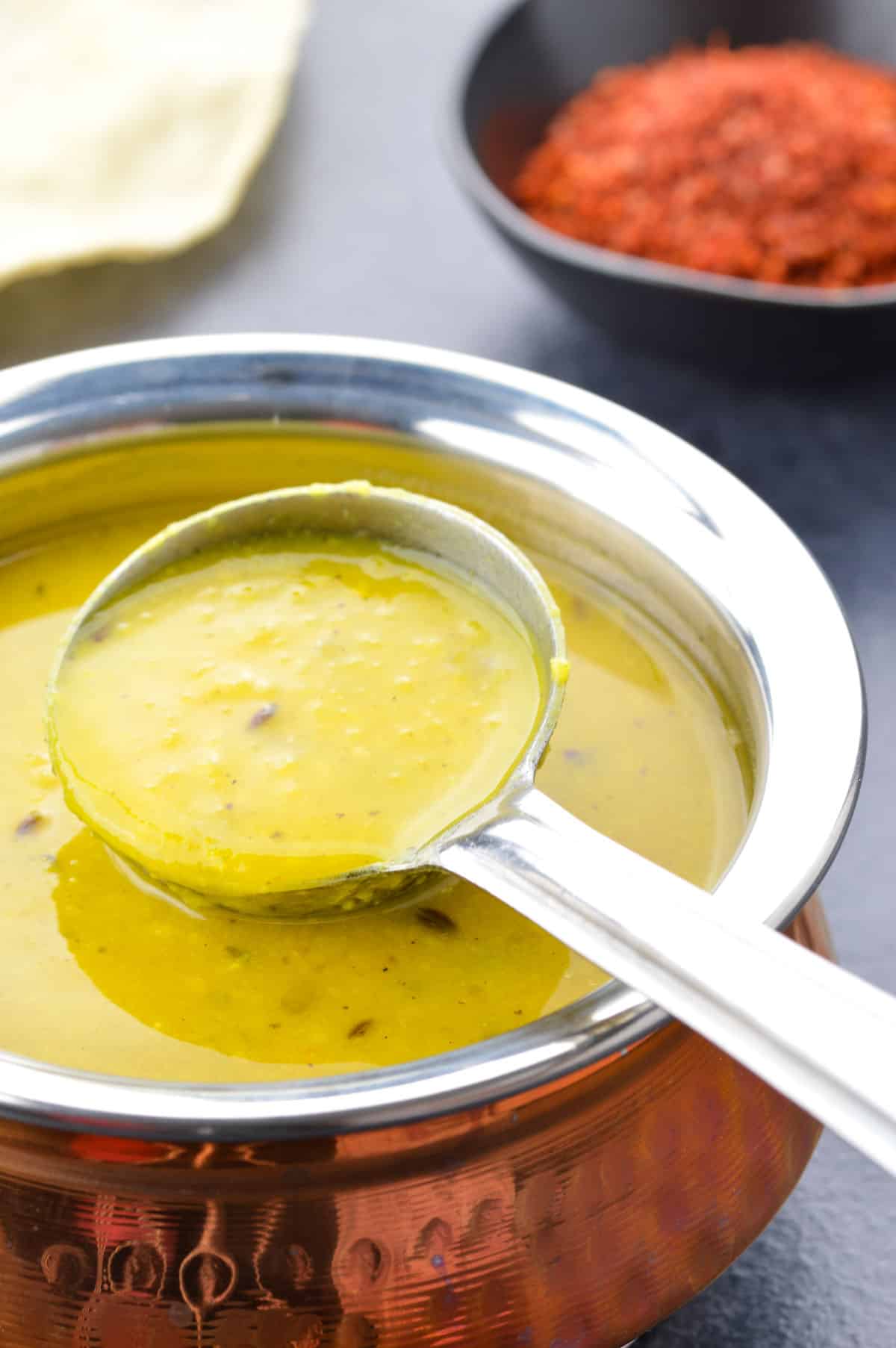 close up of a pot of dal, and a ladle taking some dal from the pot, with a small bowl of chutney and a papad in the background.
