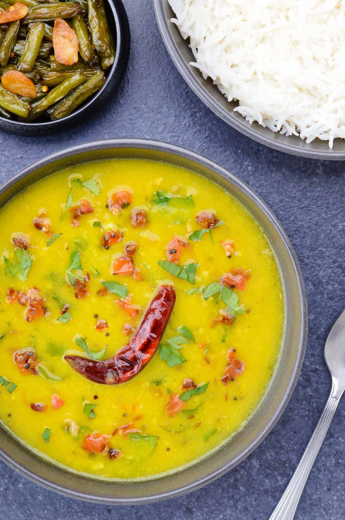 a big bowl full of homestyle dal fry, with a bowl of rice, and a small bowl of green beans stir fry on the side.