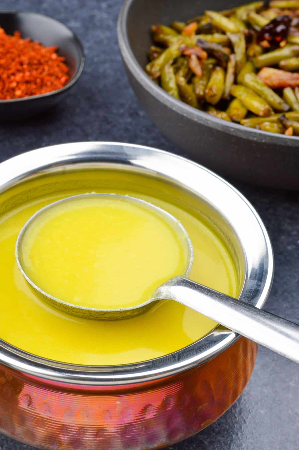 close up of a pot of yellow dal, and a ladle taking some dal from the pot, with a bowl of beans stir fry and red chutney in the background.