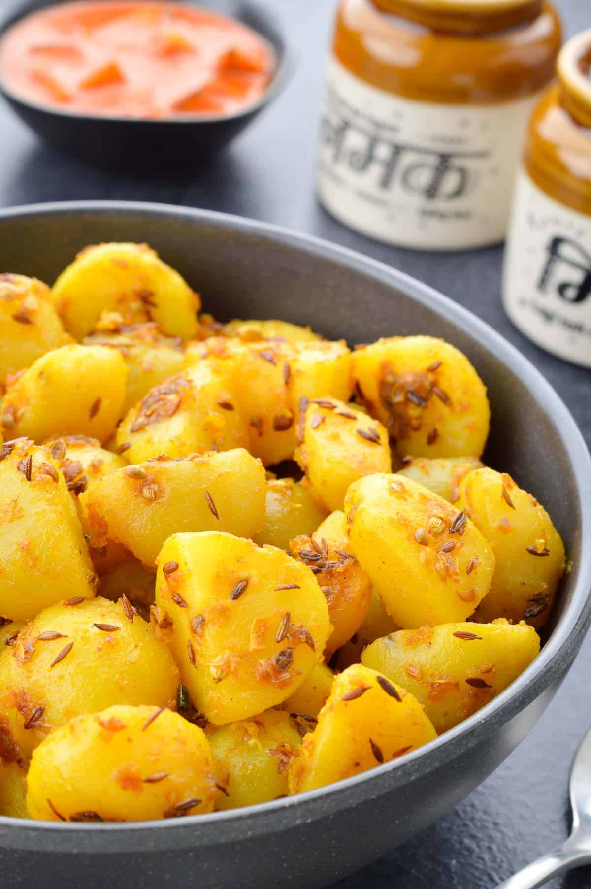 a close up shot of a bowl full of jeera aloo, with a spoon on the side, and a small bowl of pickle in the background.