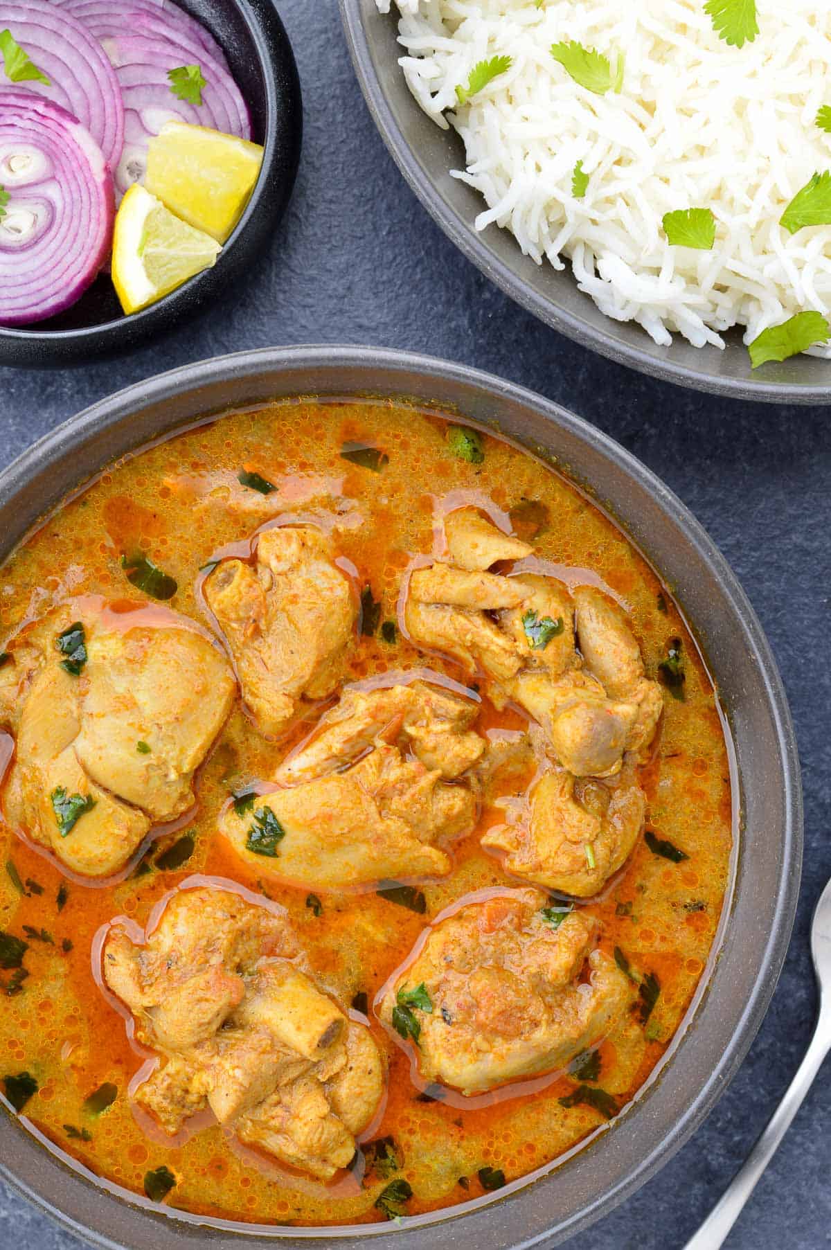 top shot of a bowl full of indian chicken curry, along with a bowl of rice, and a small bowl of sliced onions & lemon on the side.