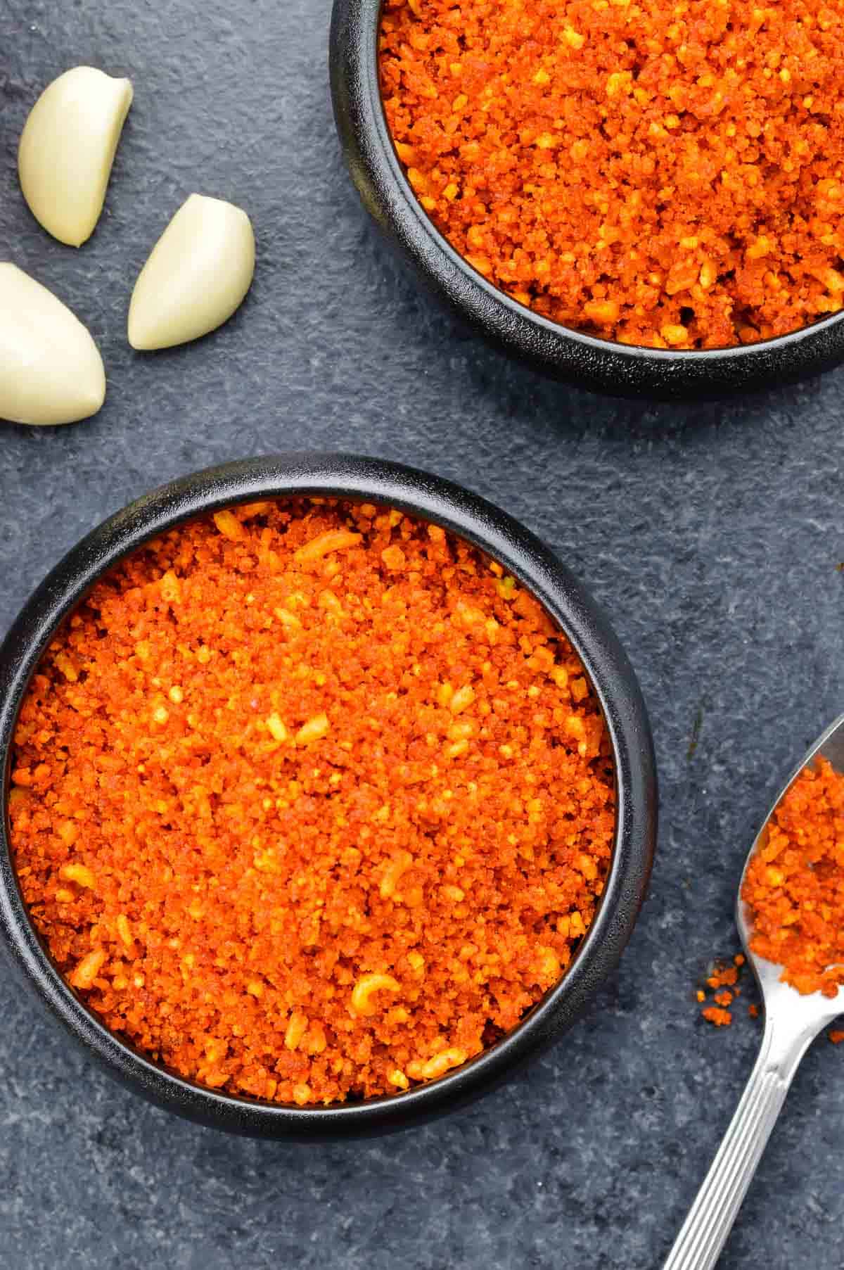 close up shot of a small bowl full of dry garlic chutney, with another bowl of garlic chutney, and some garlic cloves in the background.