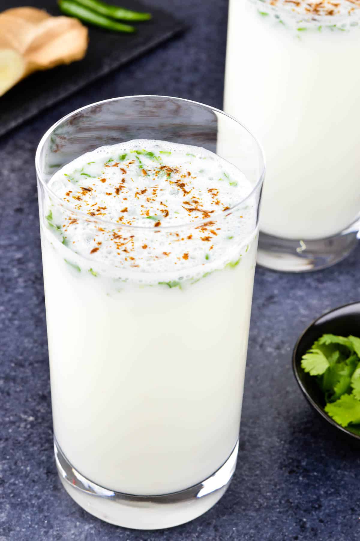 close up shot of a glass full of mattha taak, with another glass of mattha in the background, along with some whole ginger, chillies, and herbs on the side.