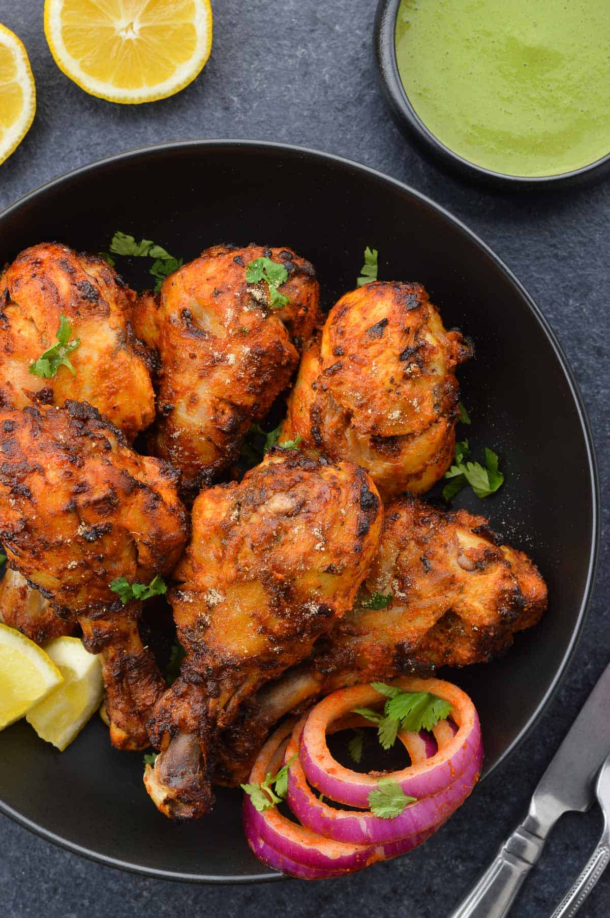 air fried tandoori chicken drumsticks in a bowl, along with laccha pyaaz, a couple lemon wedges & chopped coriander leaves, and a bowl of green chutney on the side.